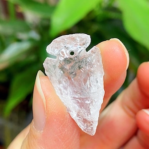 以下が含まれることがあります： 小さな穴が開いた、矢じり型の透明なクリスタルペンダント。クリスタルは質感のある表面で、ぼやけた緑色の背景を背景に指で持たれています。