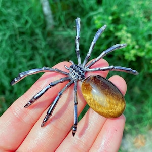 May include: A silver-toned spider-shaped pendant with black accents on the body and legs. The spider has a large, oval-shaped, tiger's eye stone as its abdomen. The pendant is displayed against a green background.