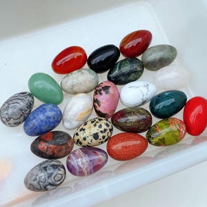 May include: A collection of twenty-one polished, egg-shaped stones in various colours and patterns, including solid red, black, green, blue, and white, as well as speckled, marbled, and banded varieties, all arranged together in a white plastic container.