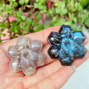 Könnte beinhalten: Zwei geschnitzte Bienenfiguren aus Stein in Form von hexagonalen Waben. Eine Figur besteht aus einem durchscheinenden, blass graubraunen Stein, während die andere aus einem dunklen, irisierenden Labradorit mit leuchtend blauen Reflexen besteht.