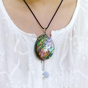 Peut inclure: Un pendentif en forme de goutte en coquille d'ormeau présentant des tourbillons irisés de vert, de violet et de bleu. Le pendentif est suspendu à un simple collier en cordon noir, mettant en valeur la surface naturelle, polie et multicolore.