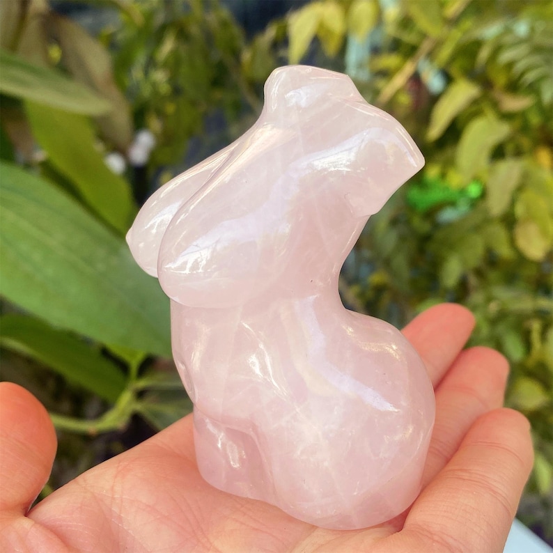 May include: A pink rose quartz torso sculpture. The sculpture is smooth and polished, with rounded forms representing the upper body. The stone has a translucent quality, with variations in the pink hue. The sculpture is held in a hand, with a green leafy background.