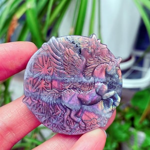 May include: A round, carved gemstone pendant featuring a winged horse, in shades of purple, pink, and gray. The pendant has intricate details, including the horse's wings, mane, and body. The background is blurred, with green foliage visible.