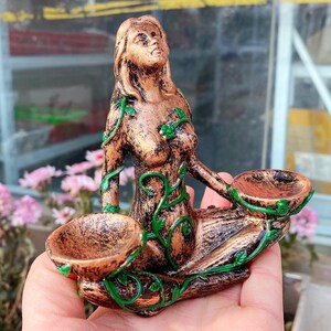May include: A bronze-colored figurine of a woman with long hair, adorned with green vine-like accents. The figure holds two small, bowl-shaped containers. The statue is likely a decorative item or a holder for small objects.