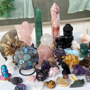 Pode incluir: Uma cole&ccedil;&atilde;o de v&aacute;rias esculturas e esculturas de pedras preciosas. Inclui um anjo rosa, um Buda preto, uma coruja verde, uma esfera de labradorita e v&aacute;rias torres de cristal. A paleta de cores &eacute; diversificada, com tons rosa, verde, preto e iridescente.