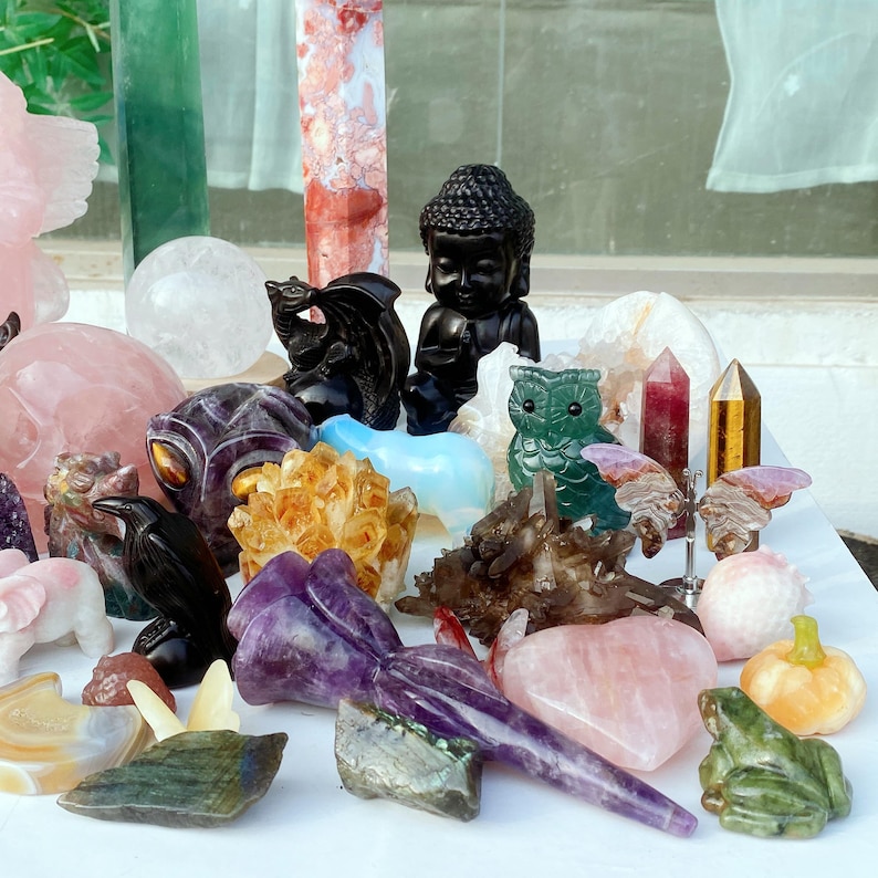 Pode incluir: Uma cole&ccedil;&atilde;o de estatuetas esculpidas em pedras preciosas, incluindo uma est&aacute;tua de Buda negra, um drag&atilde;o, uma coruja e um sapo. Outros itens incluem cristais de quartzo, um cora&ccedil;&atilde;o de quartzo rosa e uma ab&oacute;bora. A paleta de cores inclui preto, verde, roxo, rosa e dourado.