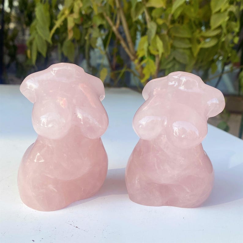 May include: Two pink rose quartz torso sculptures. The sculptures are carved in a stylized form, depicting the upper body. The smooth, polished surface reflects light, highlighting the natural variations in the stone. The sculptures are displayed on a white surface.