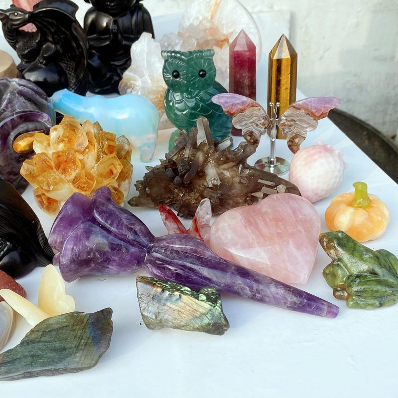 Pode incluir: Uma cole&ccedil;&atilde;o de estatuetas de pedras preciosas esculpidas, incluindo uma flor de ametista roxa, um cora&ccedil;&atilde;o rosa, um sapo verde e uma pequena ab&oacute;bora laranja. Outros itens incluem uma coruja verde, uma borboleta e pontas de cristal.