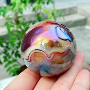 Puede incluir: Una piedra esférica pulida con un patrón de remolinos en tonos burdeos intenso, crema y azul. La superficie tiene un brillo iridiscente y reflectante que capta la luz, revelando intrincadas bandas onduladas y capas translúcidas en toda la pieza.