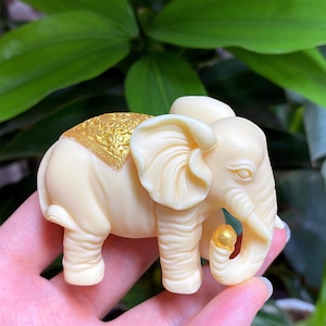 Op de afbeelding: Ivoor kleurige olifantenbeeldje met gouden accenten. De olifant heeft gedetailleerde kenmerken, waaronder grote oren, een slurf en een decoratieve gouden deken. Een kleine gouden bal wordt in de slurf gehouden. Het beeldje wordt in een hand gehouden.