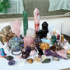 Pode incluir: Uma cole&ccedil;&atilde;o de v&aacute;rios cristais e figuras esculpidas. O sortido inclui um obelisco verde, um anjo rosa, uma est&aacute;tua de Buda preta e uma variedade de figuras de animais, esferas e outras formas em tons de rosa, roxo e transparente.