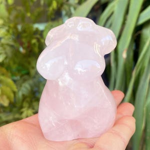 May include: A pink rose quartz torso sculpture. The sculpture is smooth and polished, with rounded forms representing the upper body. The colour is a soft, translucent pink. The sculpture is held in a hand, with green foliage in the background.