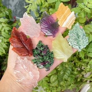 Puede incluir: Una colección de hojas talladas en piedras preciosas de varios colores, incluyendo rosa, morado, amarillo, verde y rojo. Las hojas se sostienen en una mano, mostrando las diferentes texturas y tonalidades de las piedras.