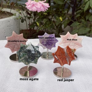 May include: A collection of gemstone sunburst sculptures, each mounted on a silver stand. The stones include strawberry quartz, dream amethyst, pink oboe, moss agate, and red jasper. The text labels each stone's name. A pink flower is in the background.