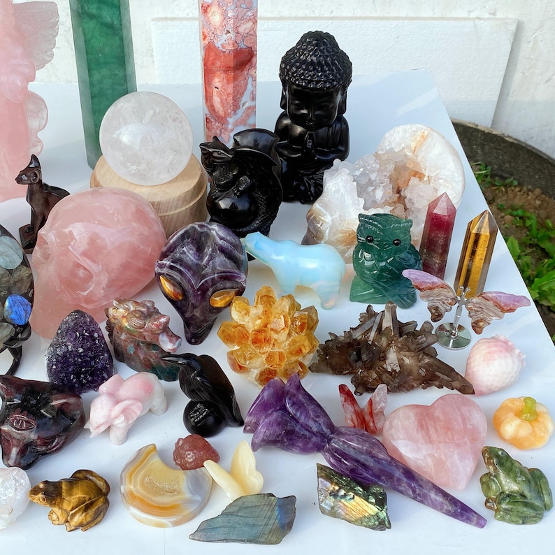Pode incluir: Uma cole&ccedil;&atilde;o de v&aacute;rias estatuetas de cristal esculpidas e pedras preciosas. Inclui uma est&aacute;tua de Buda preta, uma caveira rosa, um urso azul, uma coruja verde e uma esfera de quartzo transparente. Outros itens incluem uma borboleta, cora&ccedil;&otilde;es e pontas de cristal.