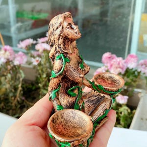 May include: A bronze-colored figurine of a woman with long hair, adorned with green vine accents. The figure holds two small, round bowls. The statue is held in a hand, with pink flowers and a window in the background.