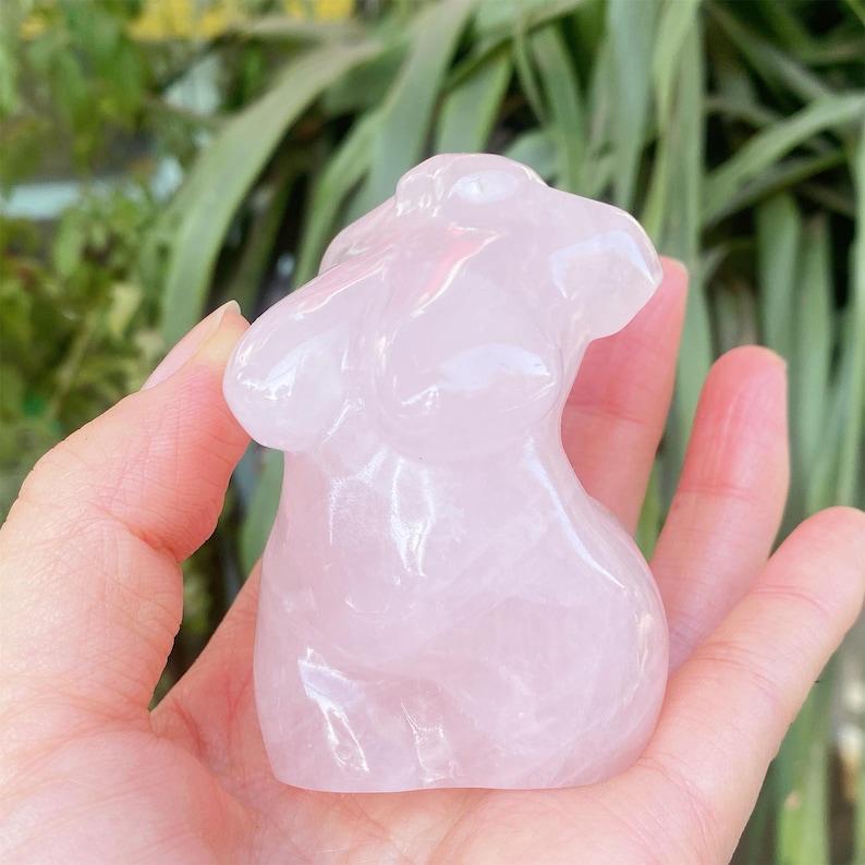 May include: A light pink rose quartz crystal carving of a torso. The sculpture is held in a hand, with a blurred green background. The carving is smooth and polished, with visible details of the form.