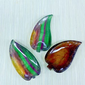 Puede incluir: Tres piedras decorativas con forma de hoja. Las piedras son translúcidas y presentan una variedad de colores, incluyendo verde, morado, amarillo y naranja. Las hojas tienen una superficie lisa y pulida y un pequeño detalle de tallo en la base. Las piedras están dispuestas sobre una superficie clara.