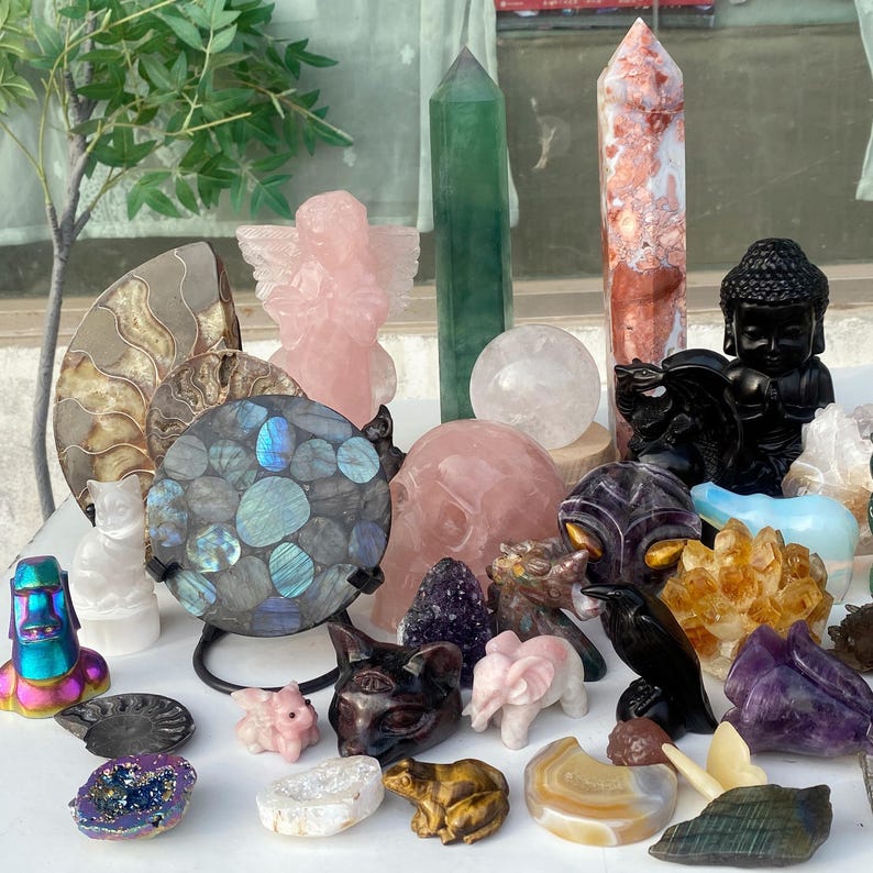 Pode incluir: Uma cole&ccedil;&atilde;o de v&aacute;rios cristais e estatuetas esculpidas. Os itens incluem um anjo rosa, um corvo preto, uma est&aacute;tua de Buda, uma esfera de labradorita e v&aacute;rias torres de cristal. A paleta de cores inclui tons rosa, verde, preto e iridescente.