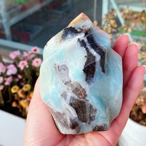 May include: A polished amazonite gemstone in a freeform shape. The stone displays a blend of light blue, brown, and white hues. The amazonite is held in a hand, highlighting its natural form and texture. The gemstone is a decorative item.