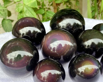 May include: A collection of polished obsidian spheres. The spheres have a dark, reflective surface with iridescent flashes of color, primarily in shades of purple, gold, and green. The spheres are arranged on a white surface, with green foliage in the background.