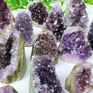 May include: A collection of amethyst crystal clusters in various sizes and shapes. The crystals display a range of purple hues, from light lavender to deep violet, with a rough, natural exterior. The clusters are arranged on a white surface.
