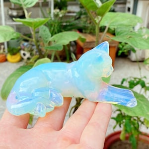 Puede incluir: Una figura de gato de opalita azul claro translúcida tallada en posición reclinada. La superficie lisa y pulida de la piedra refleja la luz con reflejos iridiscentes, mostrando la forma felina con orejas, cola y patas delanteras extendidas.