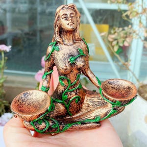 May include: A bronze-colored figurine of a seated woman with green vine accents. The woman holds two shallow bowls. The figurine has a weathered appearance and is likely made of resin or a similar material. The woman's eyes are closed.