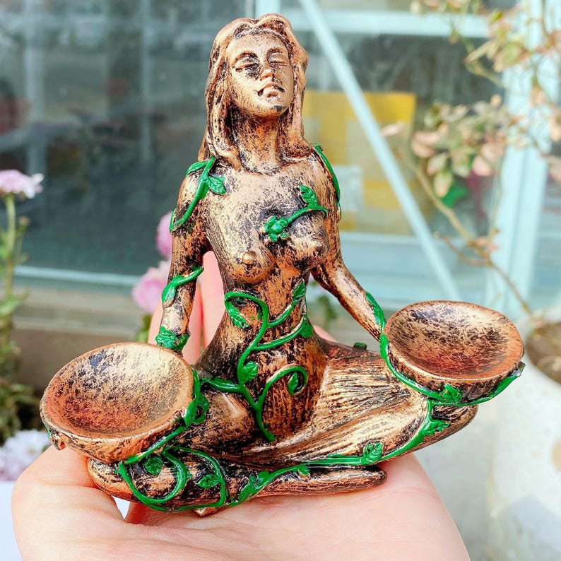 May include: A bronze-colored figurine of a seated woman with green vine accents. The woman holds two shallow bowls. The figurine has a weathered appearance and is likely made of resin or a similar material. The woman's eyes are closed.