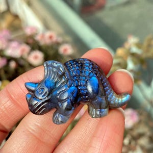 May include: A carved blue and black gemstone figurine of a Triceratops dinosaur. The sculpture has detailed features, including horns, a frill, and textured skin. The gemstone has a shimmering, iridescent quality.