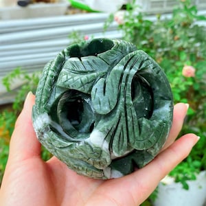 May include: A carved green stone sphere with detailed leaf and floral patterns. The sphere is held in a hand, highlighting the craftsmanship and natural colour variations. The sphere is a decorative item, likely for display.