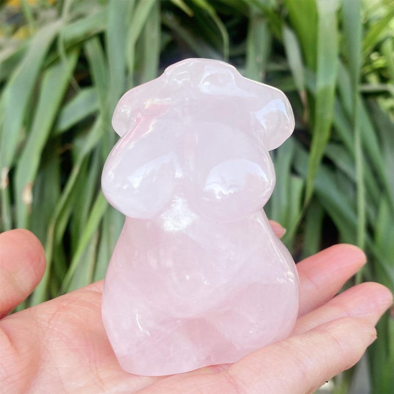 May include: A pink rose quartz torso figurine. The sculpture has smooth, rounded features and a polished surface. The figurine is held in a hand against a backdrop of green foliage.