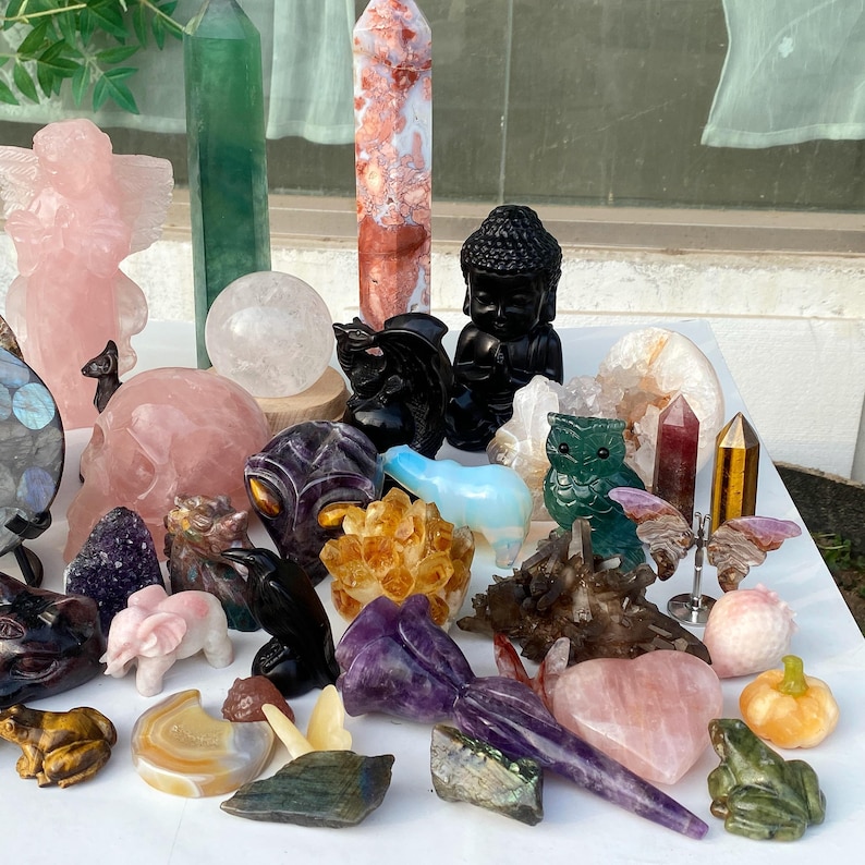 Pode incluir: Uma cole&ccedil;&atilde;o de v&aacute;rios cristais e figuras esculpidas. Os itens incluem um anjo rosa, uma caveira, uma est&aacute;tua de Buda, um sapo, uma coruja e v&aacute;rias torres de cristal. As cores incluem rosa, verde, preto e dourado.