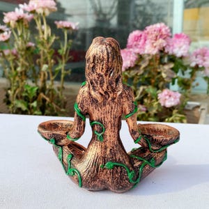 May include: A bronze-colored sculpture of a seated figure with a tree-like texture, holding two small bowls. Green vines wrap around the figure. The sculpture is likely a decorative item or candle holder.