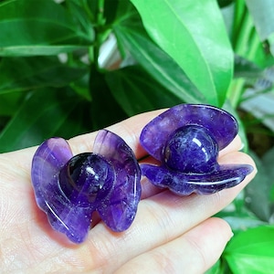 May include: Two polished amethyst crystal sculptures, each shaped like a hat with a rounded top and a flared brim. The sculptures are a deep purple color and are held in a hand, with green foliage in the background. The amethyst has a glossy finish.