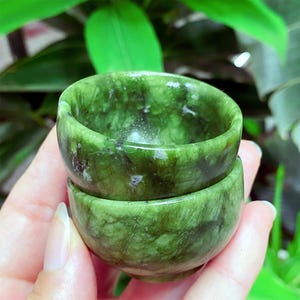 Puede incluir: Dos pequeñas tazas redondas de jade verde apiladas una encima de la otra. Las tazas tienen una superficie lisa y pulida con patrones de vetas naturales. Las tazas se sostienen en la mano de una persona, con follaje verde en el fondo.