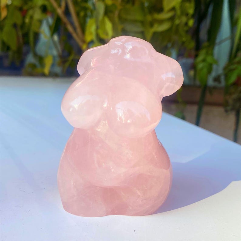 May include: A pink rose quartz torso sculpture. The sculpture is carved from a translucent, light pink stone, with smooth, rounded forms. The bust is a stylized representation of the female form, with prominent breasts and a gently curved torso. The sculpture is displayed on a white surface.