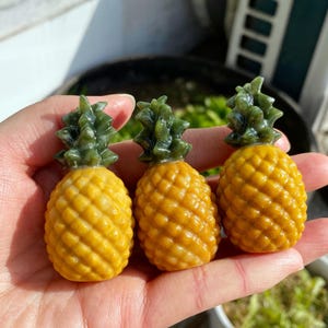 以下が含まれることがあります： 3つの小さな彫刻されたパイナップルの置物。パイナップルは、緑色のトップが付いたテクスチャ加工された黄色です。置物は人の手で持たれており、背景にはぼやけた緑があります。
