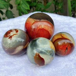 May include: Four polished stone spheres in varying sizes and colours. The largest sphere is primarily red and brown, while the smaller spheres display shades of grey, green, orange, and brown. The stones have a smooth, rounded shape.