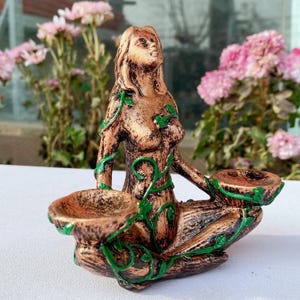 May include: A bronze-colored statue of a seated woman with green vine accents. The statue has two shallow bowls and is surrounded by pink flowers. The woman is looking upwards.