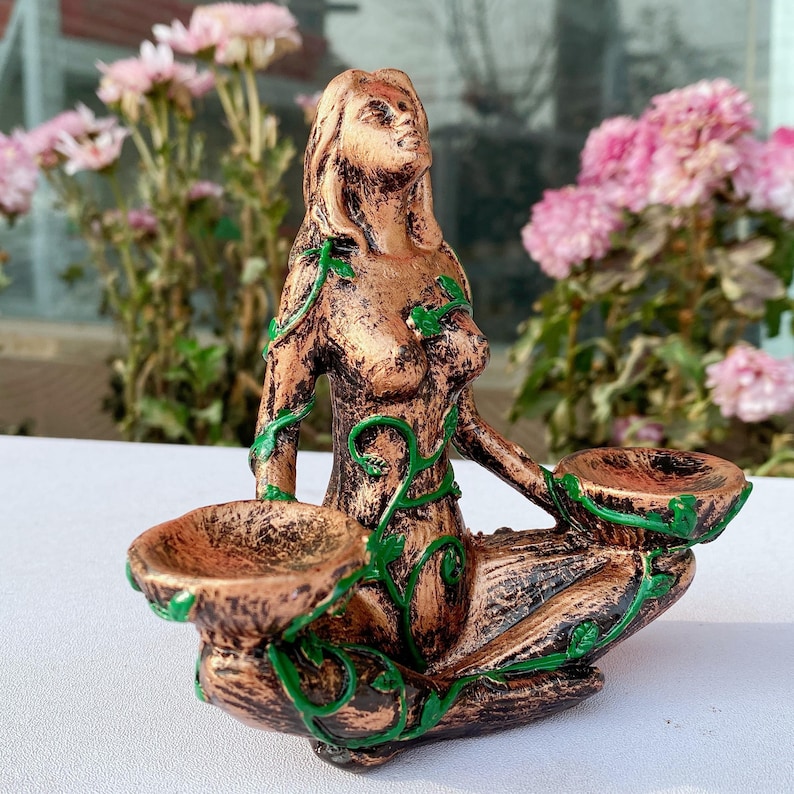 May include: A bronze-colored statue of a seated woman with green vine accents. The statue has two shallow bowls and is surrounded by pink flowers. The woman is looking upwards.