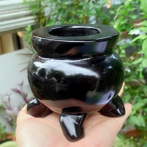 May include: A glossy black cauldron-shaped incense burner with three rounded feet. The burner has a wide opening at the top and a rounded body. The surface reflects light, highlighting its smooth texture. The object is designed for burning incense.