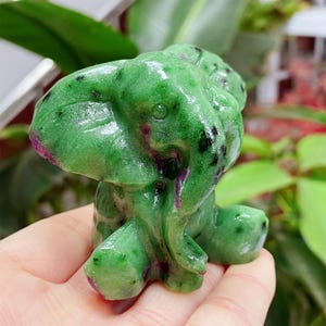 Puede incluir: Una figura de elefante de piedra preciosa verde tallada con inclusiones rosas y negras. El elefante está en posición sentada con la trompa hacia abajo. La piedra preciosa pulida tiene un acabado brillante.