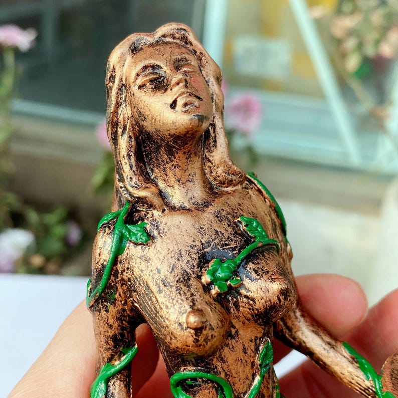 May include: A bronze-colored statue of a woman with long hair and closed eyes. Green vines with leaves are wrapped around her body. The statue has a weathered, antique appearance, suggesting it is a decorative item.