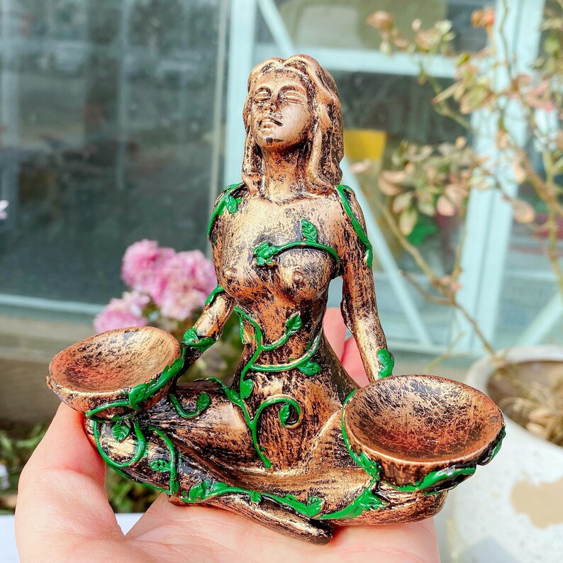 May include: A bronze-colored figurine of a seated woman with closed eyes, adorned with green vine-like accents. The figure holds two small, bowl-shaped containers. The statue is approximately 6 inches tall and is likely a decorative item.