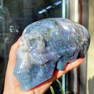 May include: A carved skull made of blue and white agate stone. The skull has detailed features, including eye sockets, a nose, and teeth. The stone has a marbled appearance with green and brown inclusions. The skull is held in a hand.