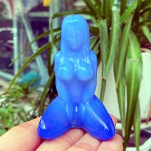 Op de afbeelding: Een doorschijnend blauw beeldje van een vrouw in knielende positie. Het beeldje heeft lang haar en is gebeeldhouwd met vloeiende rondingen. De figuur wordt in een hand gehouden, met een achtergrond van groene planten.