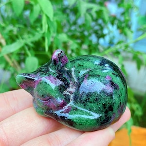 May include: A carved gemstone figurine of a sleeping cat. The cat is a mix of green and dark red colors. The figurine is held in a hand, with a blurred green background.