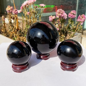 May include: Three polished black obsidian spheres of varying sizes, each on a dark red wooden stand. The spheres reflect the surroundings. The background features pink flowers and a window.