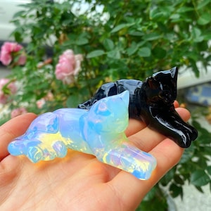 Puede incluir: Dos figuras de gato de cristal. Una es iridiscente con tonos azules, amarillos y blancos, y la otra es negra sólida. Ambas están en una pose de descanso, con las patas extendidas. El fondo presenta follaje verde y flores rosas.